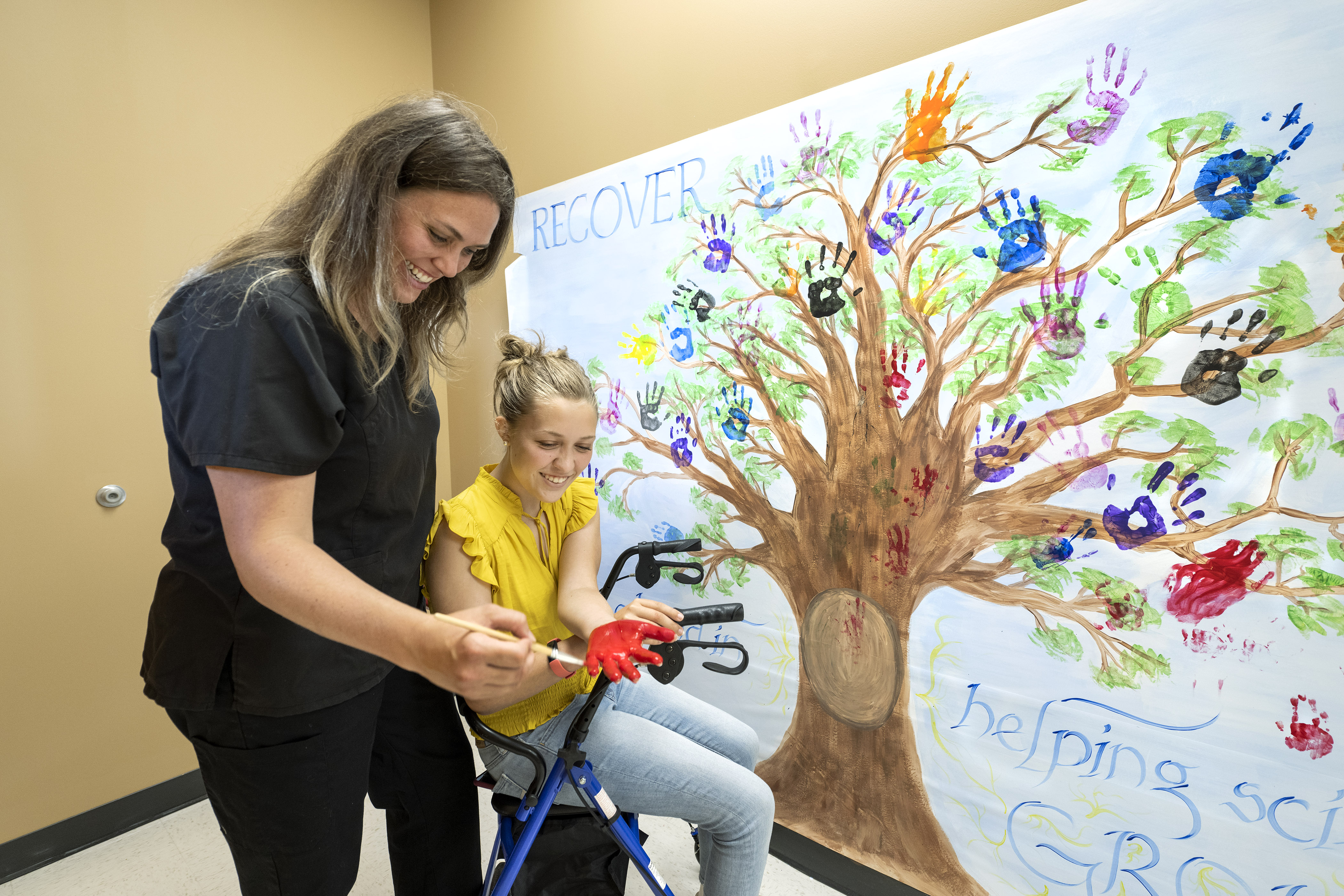 Teenage girl laughs and smiles as she puts paint on her hand. She is about to put a handprint on a wall with a tree painting and handprints as leaves. A nurse is helping her put the paint on her hand, as the girl sits on her walker.