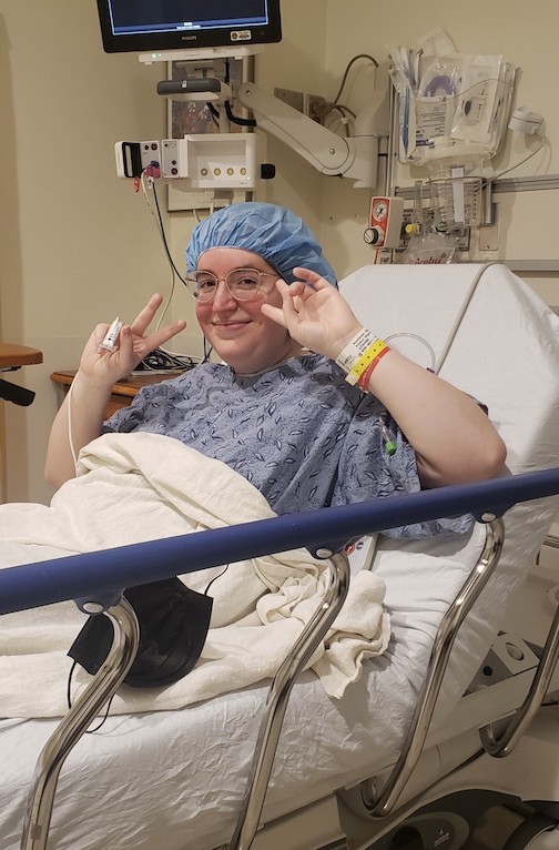 Young woman sits in a hospital bed in a hospital gown and hair net. She smiles while forming peace signs.