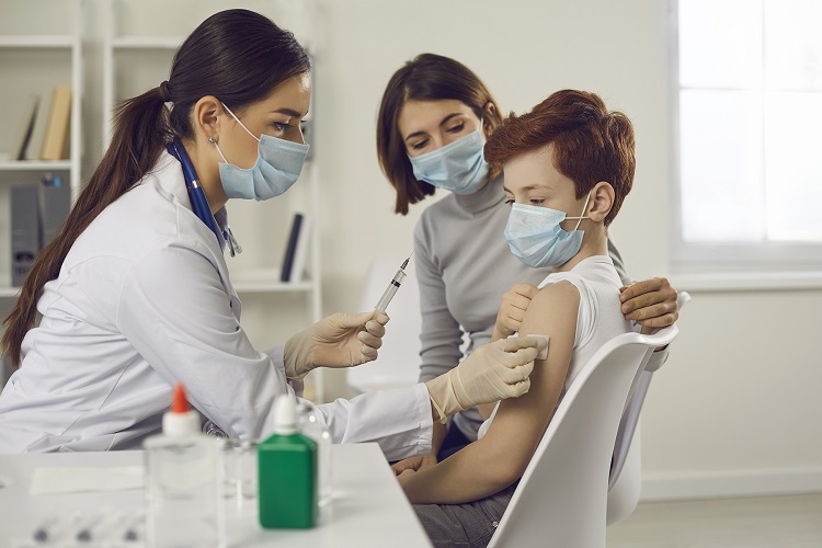 Doctor wearing a mask administers shot to young boy wearing a mask as he sits next to mother who is also wearing a mask