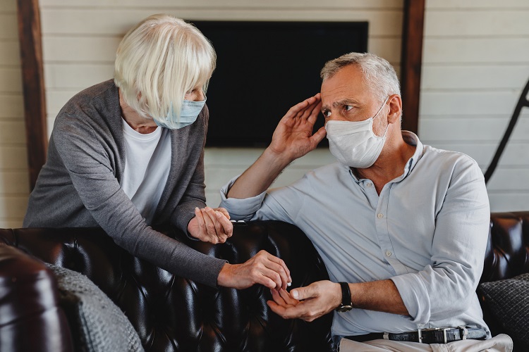 A woman hands a man a small white pill, while the man with a headache rubs his temple. Both are wearing paper face masks in their home.