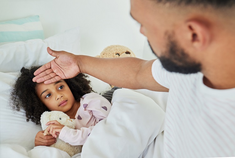 Father holding hand against forehead of young daughter lying in bed