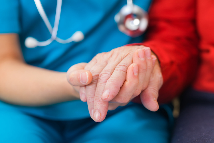A close up image of a doctor holding the hand of an older patient. The doctor’s scrubs are blue and the patient’s sweater is red.