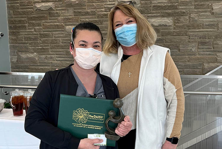 A nurse holds an award posing with a lady wearing a brown shirt and white vest