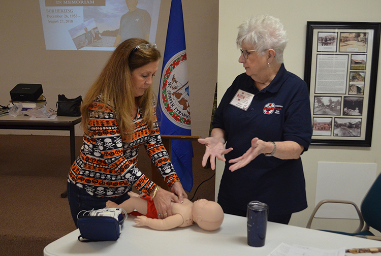 A lady practices hands-only CPR on a baby mannequin while a coach explains.