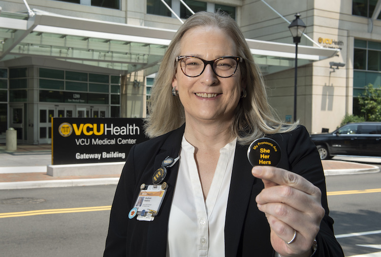 Woman stands in front of VCU Health building holding up a pin with the pronouns “she / her.”