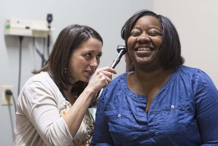 Female doctor examines the inside of a woman's ear. The doctor and woman are smiling. The doctor is wearing white and the patient is wearing a blue shirt.