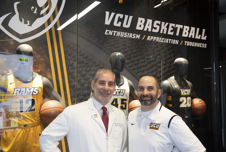 Dr. John Phillip and Jeff Collins stand together in front of VCU basketball gear. Dr. Phillips is wearing his doctor's coat while Collins is wearing VCU athletics gear.