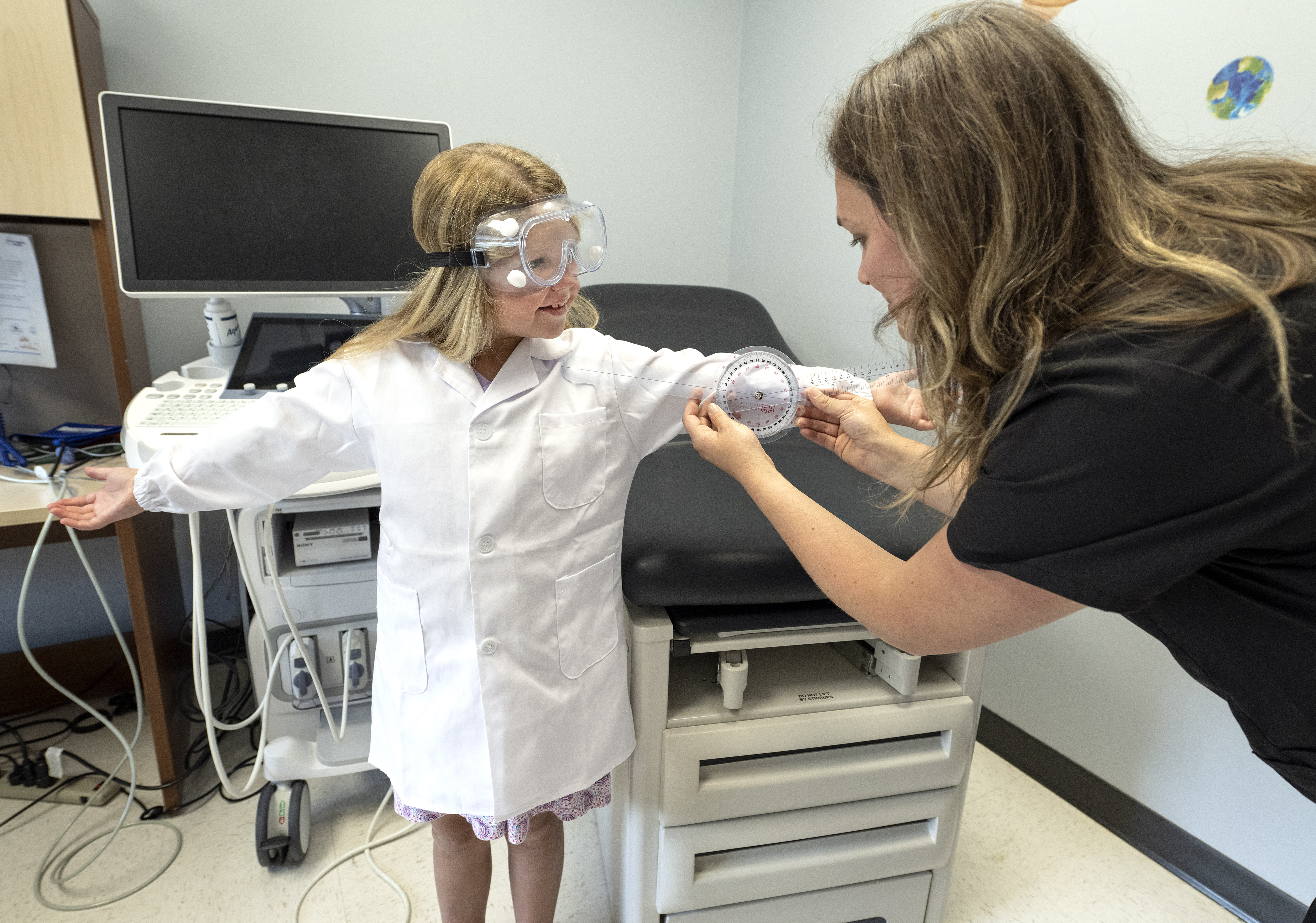 A little girl stands in a lab coat with goggles as a clinical nurse researcher measures her arms.