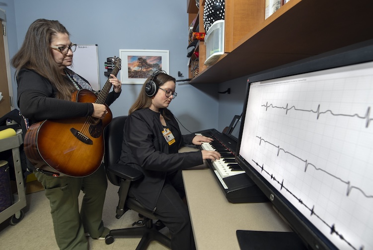 Woman plays a guitar standing next to another woman who is recording the music on a computer.