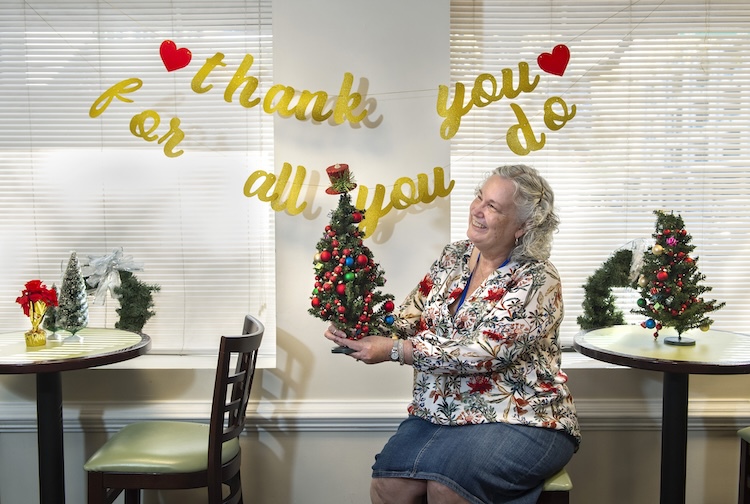 Woman sits at a table holding up a little christmas tree smiling. Behind her is a sign that says “Thank you for all you do” with hearts around it. Other tables near her also have christmas trees.