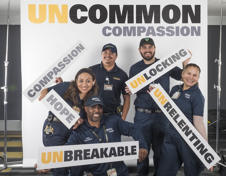 A group of team members from the hospital's facilities management department smile with signs for a photo. Signs say compassion, hope, unbreakable, unlocking and unrelenting.