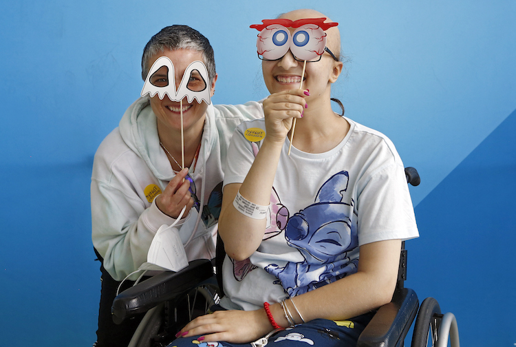An adult and teen smile with goofy masks. The teen is sitting in a wheelchair.