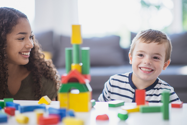 a teenager smiles at a young boy building a tower with blocks.
