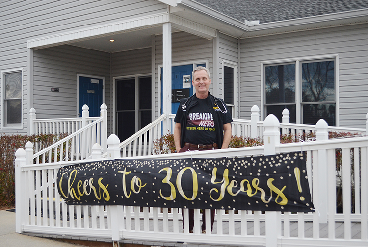 A doctor stands in front of a clinic with a banner that says, "Cheers to 30 Years!"
