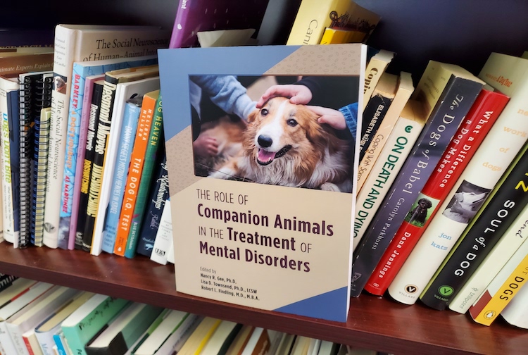 Cover of the book "The role of companion animals in the treatment of mental disorder" on a bookshelf. The book's cover as a dog on it being pet by multiple people.