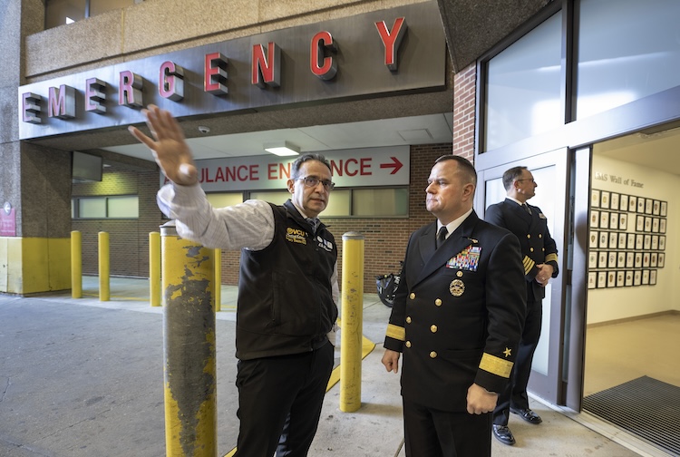 Man pointing in the other direction outside of the emergency room entrance from the street while talking to a man in a formal Navy suit.