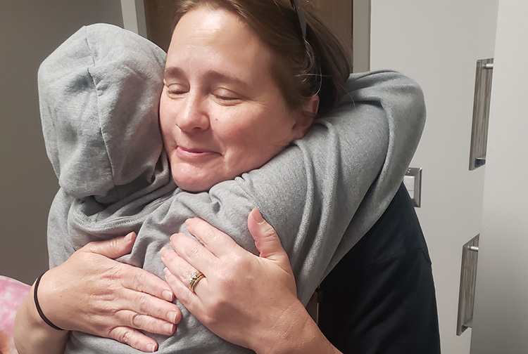 A smiling woman hugs a person wearing a hooded sweatshirt.