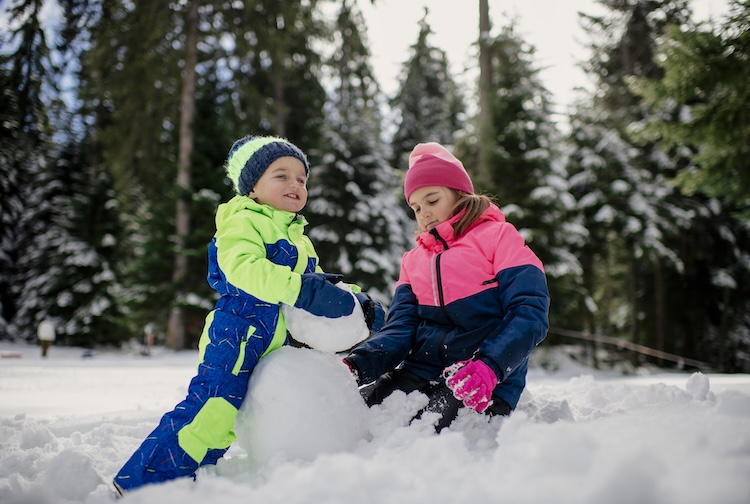 A boy and girl play in the snow. They are wearing winter jackets and are building a snowman. 