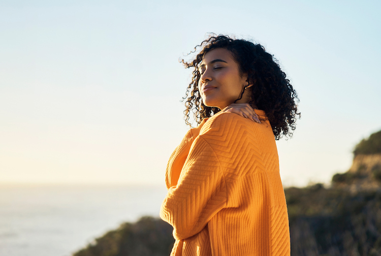 Woman stands by the sea giving herself a hug. She is smiling and wearing a comfy yellow sweater.