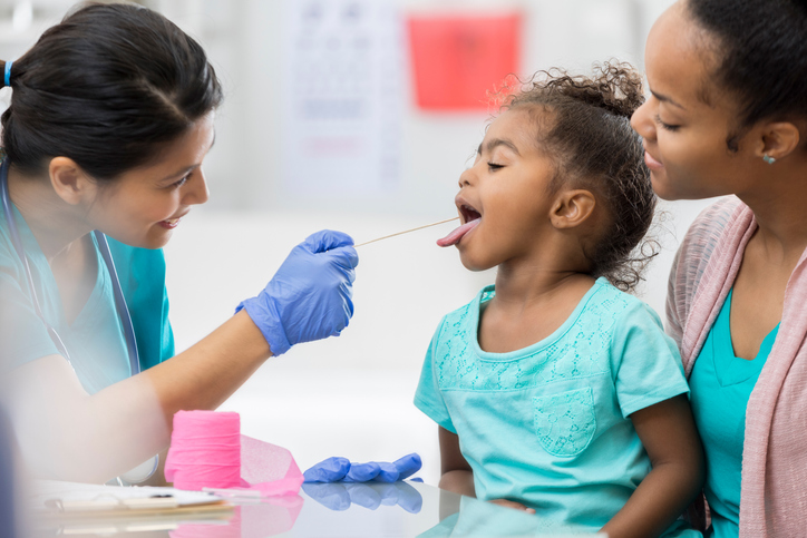 Female doctor is doing a test on a small child who is sitting on their mom's lap. The child has their tongue out, as the doctor swabs their mouth.