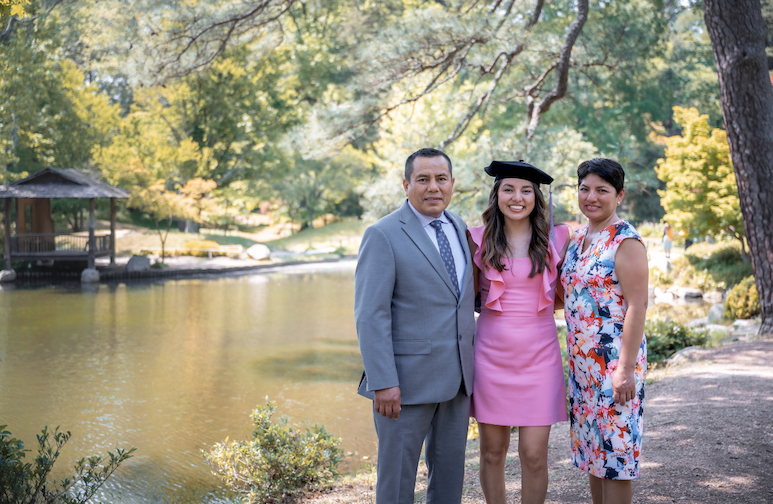 Woman with a graduation cap stands with her parents in a park for a family photo after graduation.