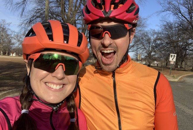 Woman and man smiling at the camera. They are wearing helmets and athletic gear for riding their bicycles. 