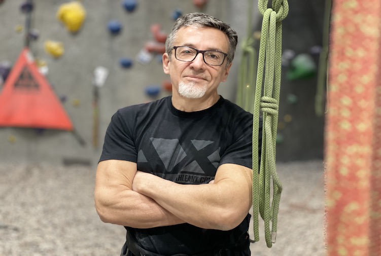Smiling, athletic man standing with arms crossed next to a climbing wall.