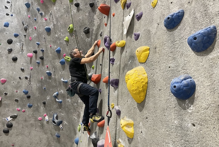 Man climbing a rock climbing wall. He is wearing a harness and is very high up from the ground.