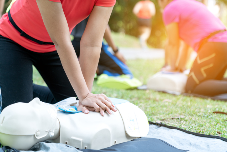 A woman participates in a first aid training class in a park by interlocking fingers over a CPR doll and giving chest compressions. 