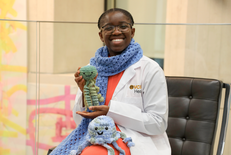 Medical student in white coat sits on a chair smiling. She is holding up her crocheted jellyfish stuffed animals. 