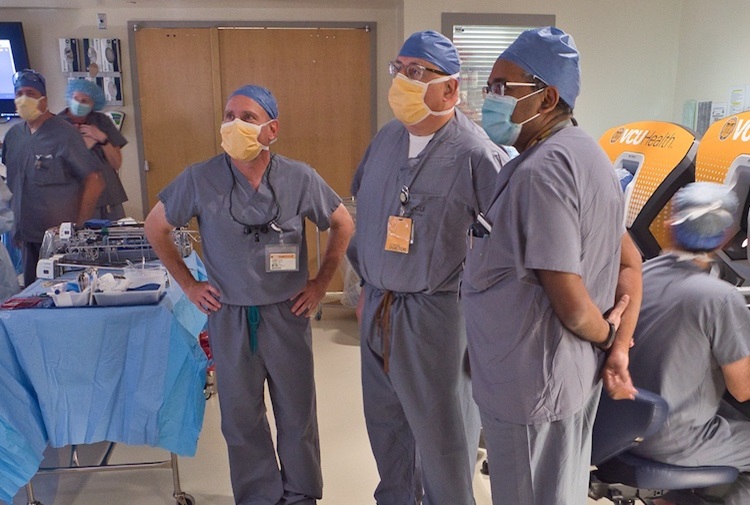 Three male doctors in blue scrubs look at the surgery being performed on a screen.