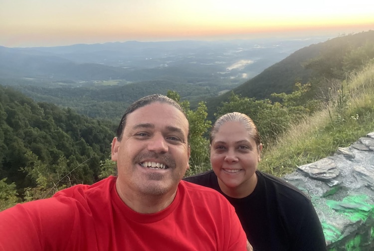  A man and woman smile while posing for a picture with mountains and a sunset behind them.