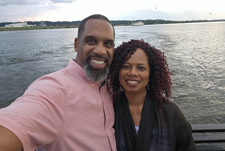 A man and woman stand together smiling with a lake behind them.