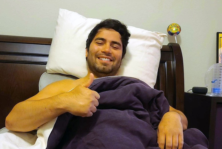 Young man smiling in a bed. He is sick, but positive. He is holding up his hand with a thumbs up.