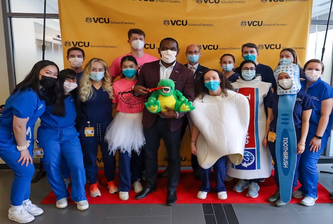 Mayor Levar Stoney stands in the middle of a large group of VCU dentistry students holding a dinosaur toy and brushing its teeth.