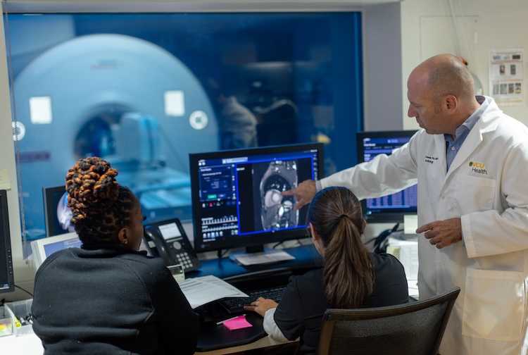 Man in a white lab coat points to MRI images on a computer. Two women in scrubs are looking at the images.