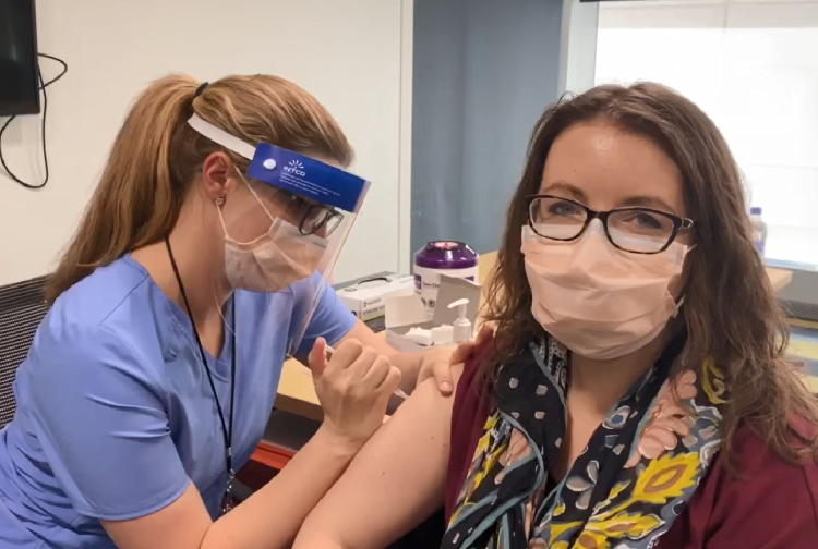 A nurse manager in blue scrubs, a face mask and a face shield injects a doctor facing the camera with a syringe full of the Pfizer COVID-19 vaccine.