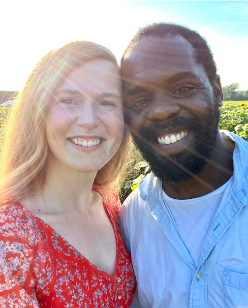 A man and woman smile on a sunny day with a field behind them.