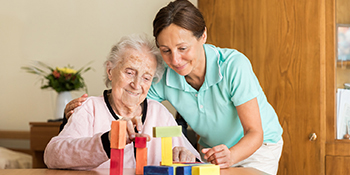 Older female patient using foam bricks on a table with another woman supervising.