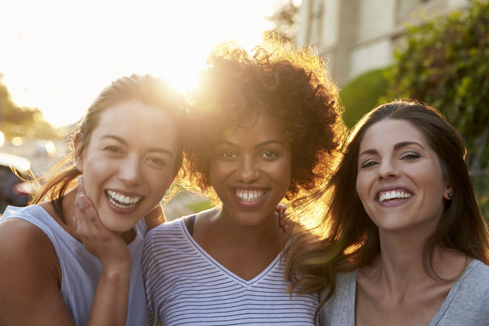 Three smiling women of different ages and ethnicities looking into the camera with the sun behind them.