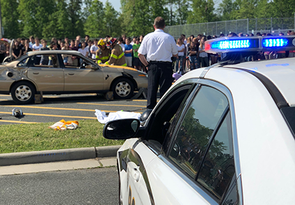 A police car in front of a car crash site, with a crowd of onlookers.