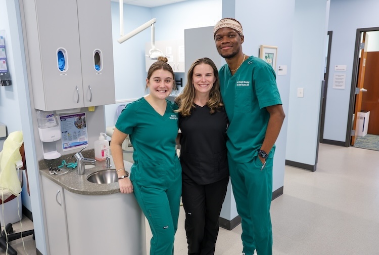 Two dental students and a dental hygienist stand together smiling in a clinic
