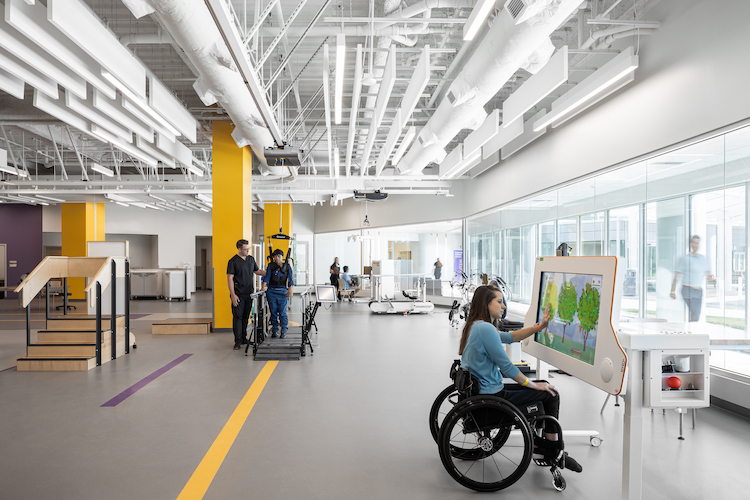 Multiple patients are in a large room using equipment to help with improving strength and mobility with their health care providers.