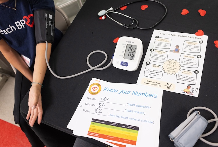 Papers on a table show blood pressure readings, with a person's arm out with a blood pressure cuff.
