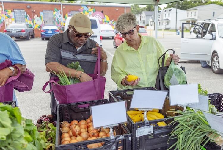 VCU Health patient is under a tent looking through crates of vegetables at a farm stand while chatting with another customer.