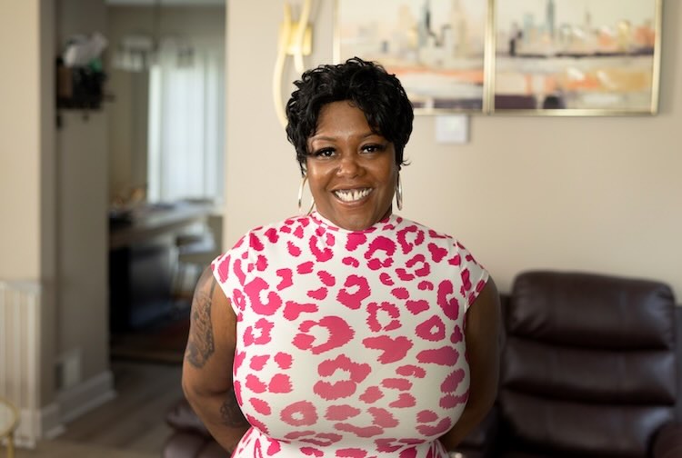 Shaneé stands in a living room wearing a white and pink shirt