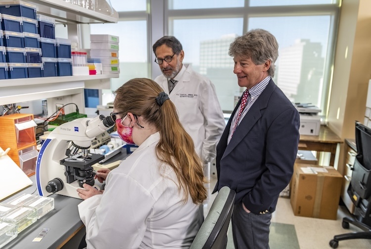 Drs. Sanyal and Stravitz stand in a research lab with a female researcher who is looking at liver cells under a microscope.