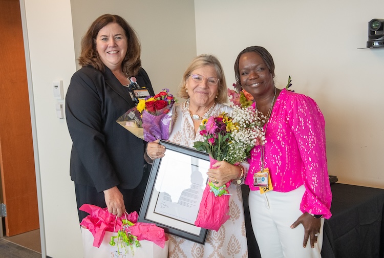 Three women stand together with a framed proclamation from the city and flowers