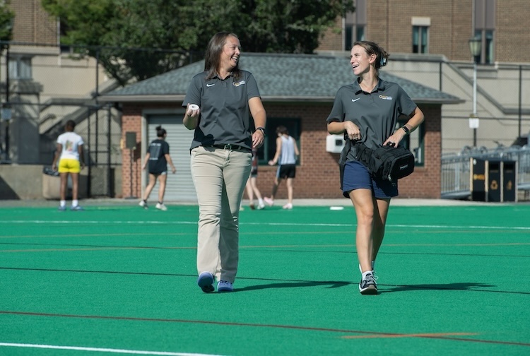 Two athletic trainers walking on a field with athletes practicing lacrosse behind them.
