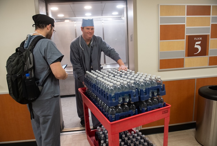 man in medical scrubs going into elevator with cart filled with water bottles
